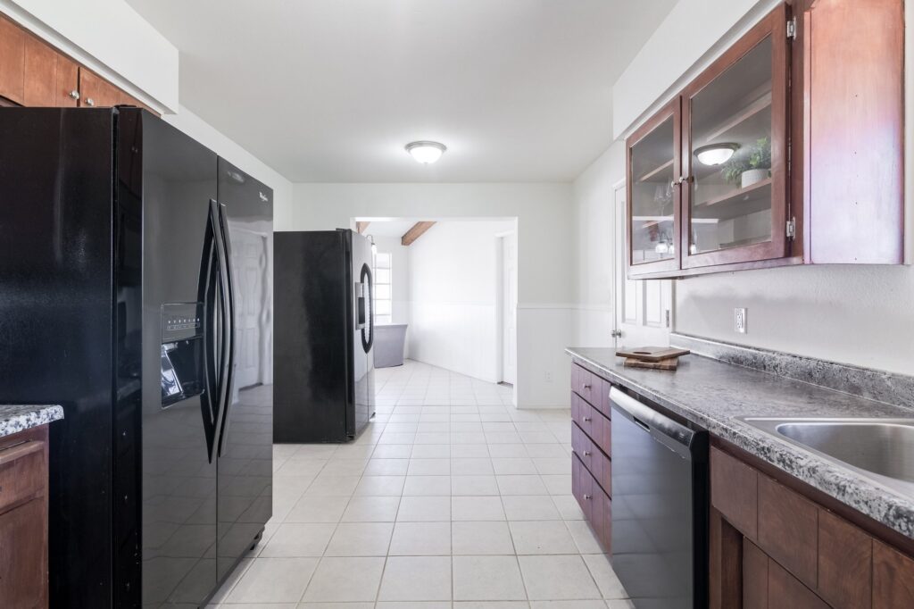 Another view of the Mesa home kitchen showing 2 matching, full-sized, black refrigerators on the left and cabinets with dishwasher on the right. Breakfast room is through a pass-through in the background.