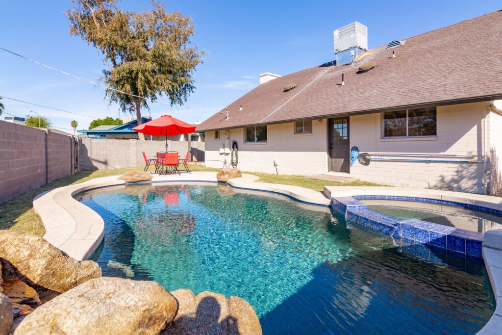 Backyard photo of PebbleTec pool and built-in spa with table and chair set at the far end and a red umbrella. Shows back side of house and lawn around pool.